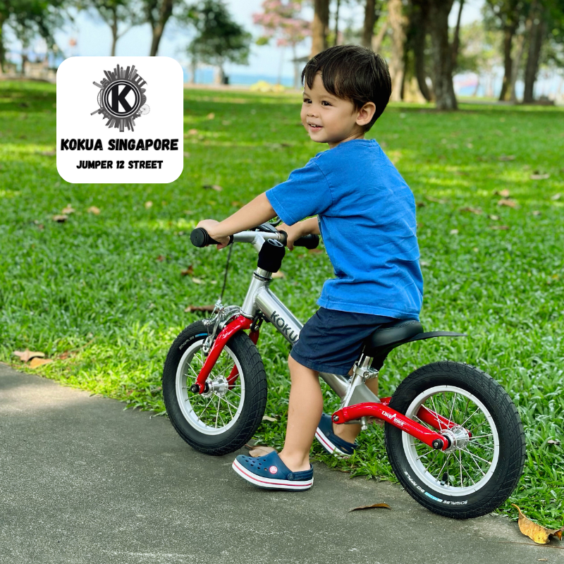 a young boy riding a red KOKUA Jumper 12 balance bike in a park