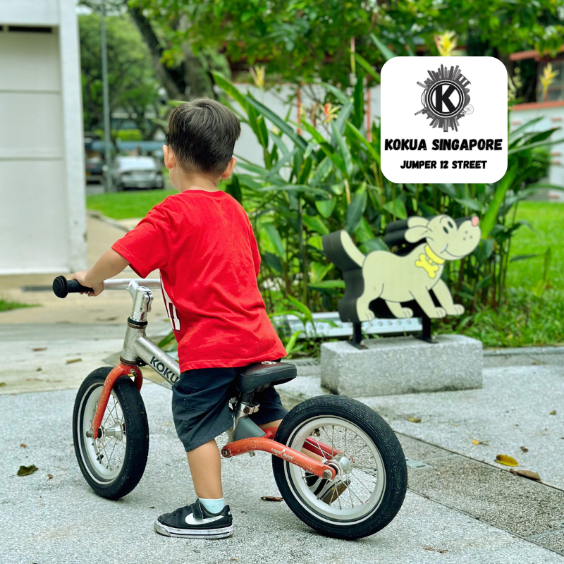 a young boy riding a red KOKUA Jumper 12 balance bike down a sidewalk