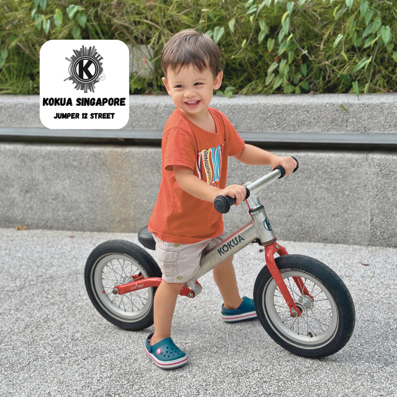 a young boy riding a red KOKUA Jumper 12 balance bike on a sidewalk