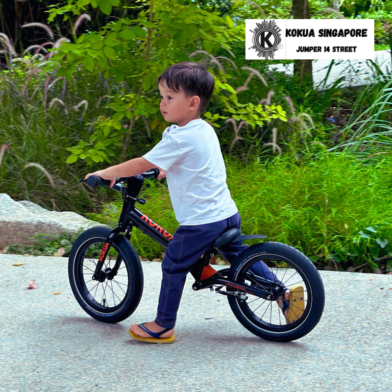 a young boy riding a black KOKUA Jumper 14 balance bike down a street